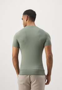 Sasniedzams īsām piedurknēm krekls olīvzaļā krāsā, izgatavots no elastīga auduma. Tam ir apaļa kakla izgriezums un gluda tekstūra ar raglanu piedurknēm.