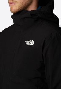 Schwarze Outdoor-Jacke mit hohem Kragen, Reißverschluss und Logo auf der linken Brust. Hergestellt aus glattem, wasserdichtem Material.