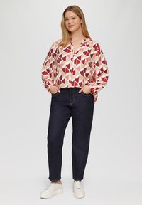 Langarmblouse met een geometrisch patroon in rood, crème en beige; gecombineerd met donkerblauwe denim broek en witte sneakers.
