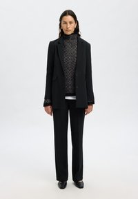 Schwarzes Blazer mit zwei vorderen Taschen, übereinander getragen mit einem gemusterten Schwarz-Weiß-Pullover mit Rollkragen, kombiniert mit schwarzen weiten Hosen und schwarzen Schuhen.