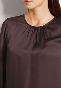 Frau mit einer dunkelbraunen Satinbluse mit rundem Ausschnitt und kleinen Falten in der Nähe des Kragens, die nur Hals und Kinn zeigt.