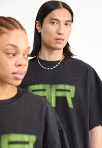 Μαύρα oversized μπλουζάκια με κοντό μανίκι και crew neck, που διαθέτουν μια πράσινη γραφική σχεδίαση "AR". Έχουν φθαρμένη υφή και είναι ορατή μια αλυσίδα από ασήμι.