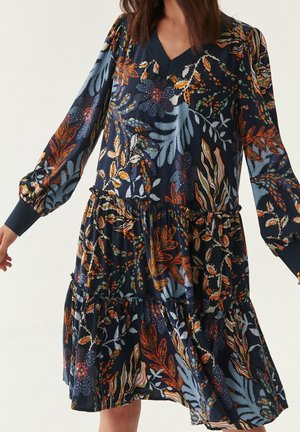 Femme portant une robe bleu marine à manches longues avec des motifs floraux et feuillus orange, blanc et vert, avec un décolleté en V et une jupe à volants.