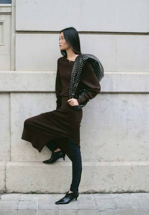 Femeie cu păr lung și întunecat, purtând o rochie închisă la culoare, colanți negri și pantofi cu toc, stând sprijinită de un zid de piatră și ținând o jachetă cu ținte.