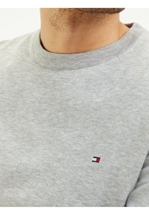 Man draagt een grijze trui met ronde hals met een klein rechthoekig logo in rood, wit en marineblauw op de borst.