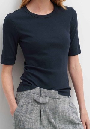 Vrouw draagt een aansluitend navy blauw kortemouwen topje gecombineerd met hooggetailleerde grijze geruite broek, staand met één hand in de zak.