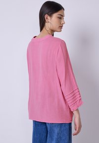 Femme aux longs cheveux noirs portant de grandes boucles d'oreilles, un haut rose texturé à manches longues et un jean bleu, se tenant de profil devant un arrière-plan uni.