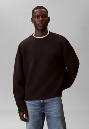 Hombre con sudadera negra de Calvin Klein y vaqueros azules, de pie frente a un fondo gris liso.