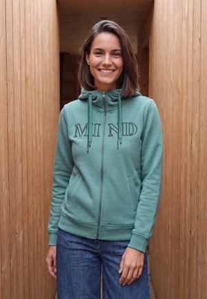 Jeune femme souriante, portant un sweat à capuche vert zippé avec le texte "MIND" et un jean bleu, debout entre des murs en bois.