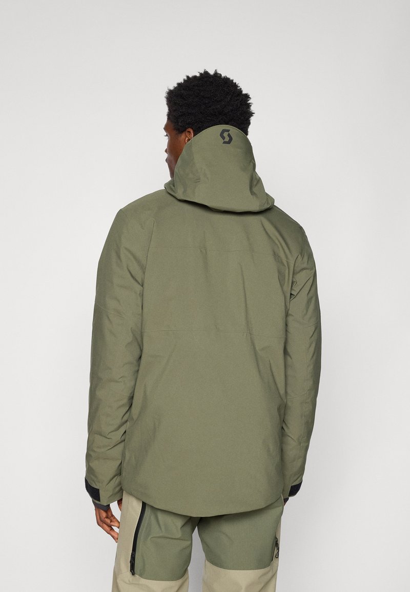 Grüne wasserdichte Jacke mit Kapuze, einschließlich eines Logos auf der Rückseite, strukturiertem Stoff und elastischen Bündchen. Getragen mit passenden hellbeigen Hosen.