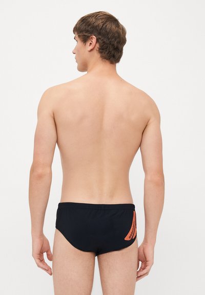 Junger Mann, der mit dem Rücken zur Kamera steht, trägt schwarze Schwimmshorts mit einem orangefarbenen Logo auf der rechten Seite vor einem schlichten weißen Hintergrund.