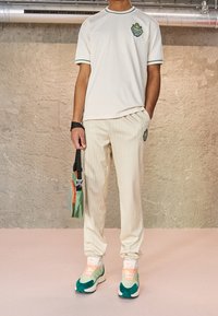 Person klädd i en krämfärgad t-shirt med korta ärmar och randiga byxor med matchande kragar, gröna och persikofärgade sneakers, hållande en liten grön och orange väska.