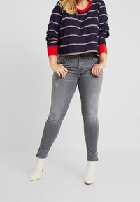 Marynarski i czerwony prążkowany krótki sweatshirt z długimi rękawami, w zestawieniu z szarymi obcisłymi dżinsami z wyblakłymi łatami, zużytym wyglądem i lekkim przetarciem.