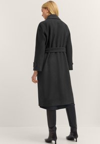 Langer schwarzer Mantel mit taillierter Taille, breitem Kragen und knöpfbaren Manschetten. Aus strukturiertem Stoff gefertigt, reicht er bis zur Mitte der Waden für eine maßgeschneiderte Passform.