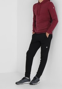 Sweat à capuche bordeaux avec une poche avant et joggers noirs avec logos. Tenue complète avec des chaussures de sport grises et une coupe décontractée.