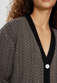 Černý a hnědý kardigan s vzorem rybí kosti, výstřihem do V a jedním bílým knoflíkem vpředu. Měkká pletená textura.