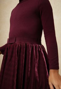 Dlhý rukáv šaty v hlbokej burgundskej farbe s priliehavým vrchným dielom a plisovanou zamatovou sukňou, s pásom z rovnakého materiálu v oblasti pása.