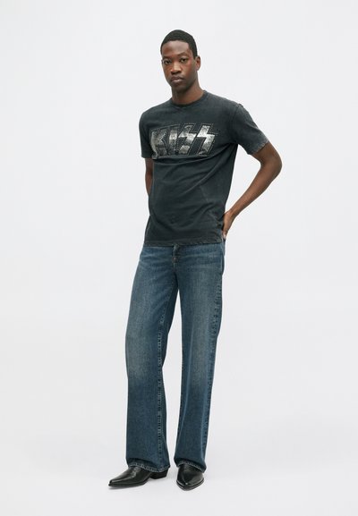 Camiseta gráfica negra con logo metálico de KISS, combinada con pantalones de mezclilla azul de pierna ancha y botas negras de punta afilada. Diseño sencillo, ajuste casual.