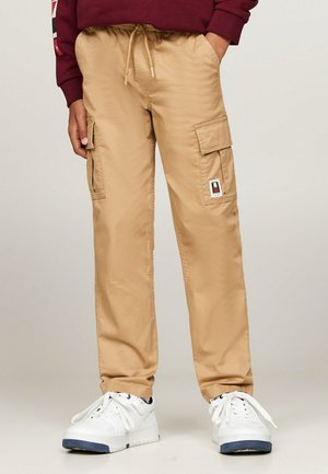Pantaloni cargo beige con vita elastica, tasche frontali e patch con logo. Indossati con sneakers bianche con accenti neri e blu.