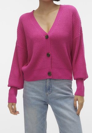 Femme portant un cardigan en maille rose vif boutonné, avec un col en V profond et des manches amples, associé à un jean bleu clair.