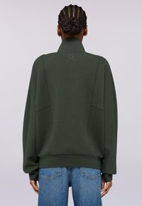 Zelená mikina s vysokým límcem, volný střih, přední kapsy a minimální detaily. V kombinaci s modrými džíny dokončuje outfit.