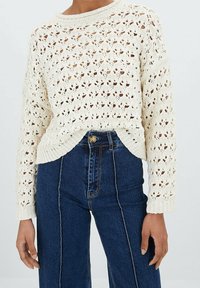 Krémově zbarvený pletený svetr s průsvitným vzorem, širokými rukávy a zaobleným výstřihem. Kombinováno s tmavými džíny s vysokým pasem.