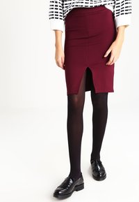 Burgundy stretchkjol med en frontslits, kombinerad med svarta ogenomskinliga strumpbyxor och glansiga svarta loafers. Toppen har ett svartvitt randigt mönster.