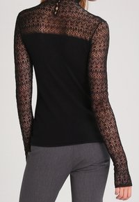 Svart långärmad topp med genomskinliga spetsärmar med intrikata mönster, nyckelhålsdetalj baktill och en figurnära silhuett.