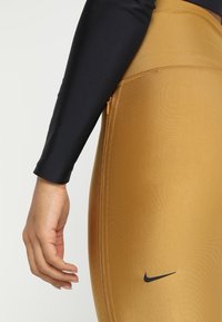 Leggings sportivi realizzati in un tessuto dorato lucido con logo nero. Presentano un'alta vita, una texture liscia e un design aderente. È visibile una manica nera lunga.