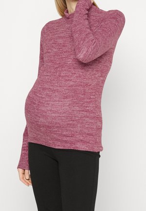 Femme enceinte portant un pull rose à manches longues et un pantalon noir, la main reposant près du visage sur fond blanc.