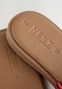 Brūna flip-flopa ar teksturētu materiālu, kuram ir iegravēts logo "NĀKAMĀS MŪŽĪGĀ KOMFORA". Ieskaitot izmēra apzīmējumus, apgriezto siksnu detaļas.
