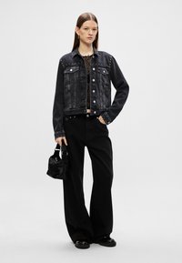 Mustat farkkutakki, jossa on niittejä, kaksi etutaskua, nappikiinnitys, yhdistettynä leveälahkeisiin mustiin housuihin ja pieneen mustaan käsilaukkuun.