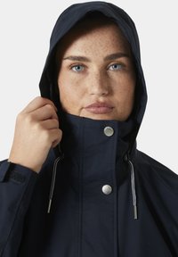 Granatowa wodoodporna kurtka z kapturem, z zatrzaskami i akcentami w postaci sznureczków. Gładki materiał o matowym wykończeniu oraz szczegóły na mankietach z rzepami.
