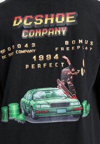 Svart t-shirt med en grafik som visar en skateboardåkare över en grön bil, omgiven av fat. Texten inkluderar "DC SHOE COMPANY" och "1994."
