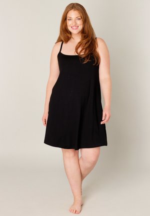 Lächelnde Frau mit langen roten Haaren, die ein ärmelloses schwarzes Kleid trägt, barfuß vor einem schlichten hellen Hintergrund steht.