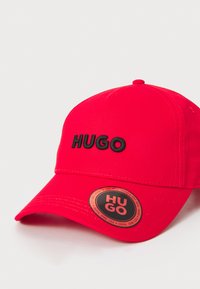 Rød baseballcap lavet af holdbart stof, med et sort broderet "HUGO" logo og et rundt mærkeskilt på siden.