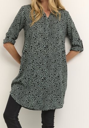 Kvinde iført grøn tunika med sort leopardprint og lange ærmer, ærmerne rullet op, kombineret med sorte leggings, stående mod en hvid væg.