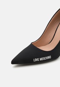 Črne salonarji z ostrim nosom, izdelani iz mehkega blaga, na katerega je ob strani v beli barvi napisan logotip "LOVE MOSCHINO". Gladka notranja obloga z rahlo teksturo.
