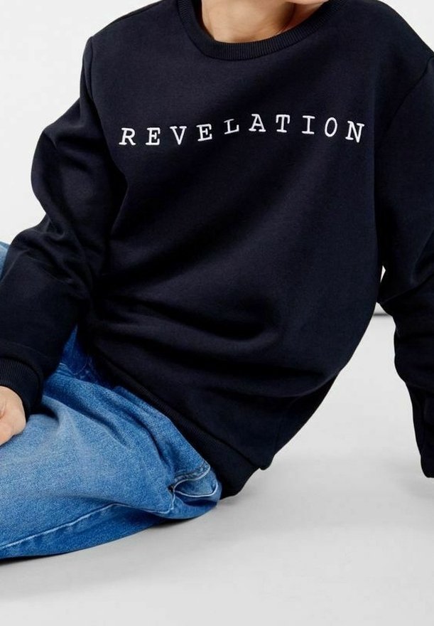 Personne portant un sweat-shirt noir avec l'inscription « REVELATION » en lettres blanches sur la poitrine, associée à un jean bleu, assise par terre.