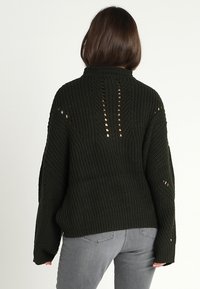 Pull en tricot vert foncé avec un col montant, présentant un motif texturé et des accents de maille ouverte le long du dos et des manches.