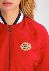 Czerwona kurtka bombowiec z prążkowanym granatowym kołnierzykiem, zamkiem błyskawicznym z przodu oraz owalną naszywką z haftem z napisem "MIŁEGO DNIA" w otoczeniu kwiatowego wzoru.