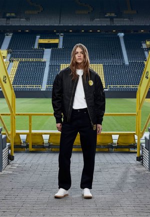 Jeune femme se tenant sur une passerelle de stade, portant une veste bomber noire avec le logo jaune BVB, une chemise blanche, un pantalon noir et des baskets blanches.