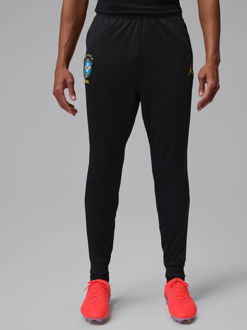 Man draagt zwarte Braziliaanse voetbalbroek met gele logo en felrode voetbalschoenen, staand tegen een grijze achtergrond.