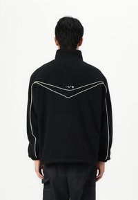 Giacca in pile nera con colletto alto, dettagli in piping bianco e una piccola grafica sul retro. Vestibilità comoda e rilassata con polsini elasticizzati.