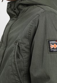 Olivově zelená bunda s vysokým límcem, zapínáním na zip, bočními kapsami a nášivkou s logem Union Jack na rukávu. Vyrobena z texturované látky.