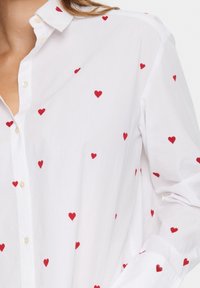 White button-up shirt made of lightweight fabric, featuring scattered red embroidered hearts. Includes a collar and cuffed sleeves.