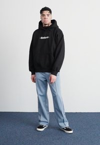 Black hoodie with white "Carhartt" logo, light blue wide-leg pants, and black sneakers with white soles, standing on a blue carpeted floor.