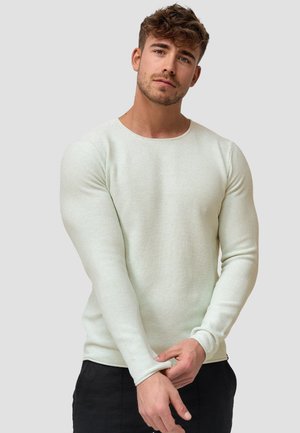 Jeune homme aux cheveux bruns courts, portant une chemise légère texturée à manches longues et un pantalon noir, ajustant sa manche devant un fond uni.