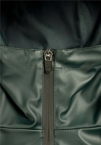 Närbild av mörkgrön slät läderplagg med en svart dragkedja som har ett litet dödskalle- och korsbenemblem på dragfliken.