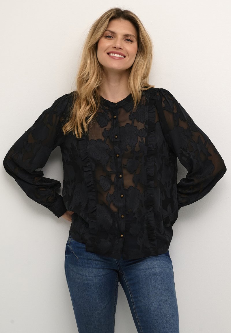Blusa nera trasparente con texture floreale, caratterizzata da bordi arricciati, un frontale con bottoni e maniche lunghe. Abbinata a jeans in denim blu.